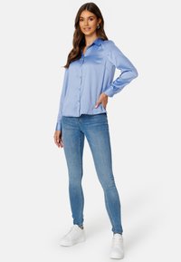 Camicia di raso blu chiaro con colletto e chiusura a bottone, abbinata a jeans blu aderenti e sneaker bianche. Design semplice e texture morbida.