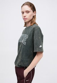 Tmavohnedé tričko s krátkym rukávom s vyblednutou textúrou, s bielym logom "NIKE" na prednej strane a malým logom na ľavom rukáve.