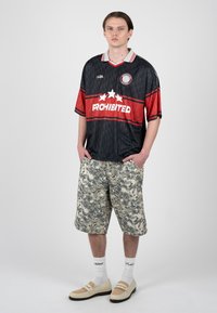 Joven con las manos en los bolsillos, vestido con una camiseta deportiva negra y roja, pantalones cortos de camuflaje, calcetines blancos y zapatos mocasines beige sobre un fondo liso.