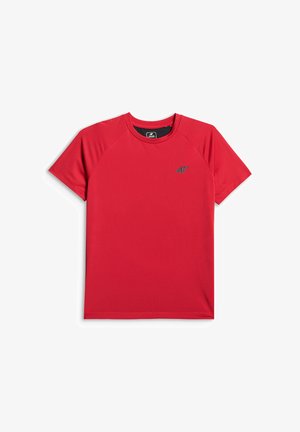 Enfant souriant debout avec une main dans la poche, portant un t-shirt rouge à manches courtes, un pantalon de sport noir et des chaussures, sur un fond neutre.