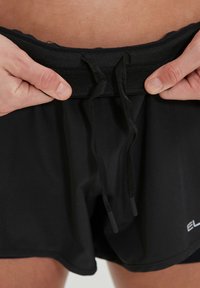 Shorts de bain noirs avec une taille à cordon, présentant une texture lisse et un logo sur le bas à droite. Vue rapprochée de la taille.