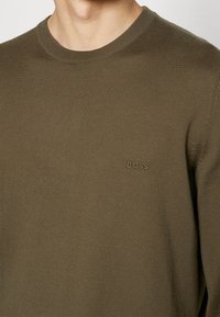 Sweter o kroju crewneck w oliwkowej zieleni, wykonany z teksturowanej dzianiny, z subtelnym tłoczonym logo na lewej piersi.