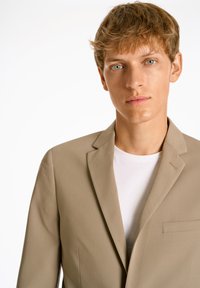 Hellbeige Blazer aus glattem Stoff, mit einer einzelnen Brusttasche, Reverskragen und einem schlichten Design. Kombiniert mit einem weißen T-Shirt.