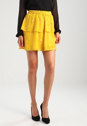Jupe jaune à plusieurs couches avec un design à volants, présentant une texture légère et des points colorés éparpillés. Taille élastique, associée à des talons noirs.