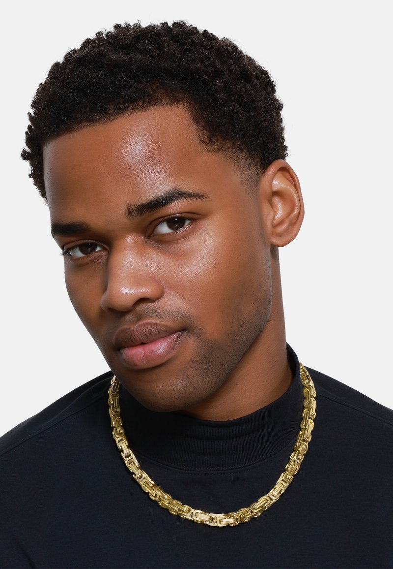 Gullkjede laget av sammenkoblede rektangulære ledd, med en polert overflate. Modell bærer en svart turtle neck.