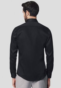 Czarna koszula z długim rękawem o gładkiej fakturze, klasycznym kołnierzyku i mankietach zapinanych na guziki. Widok z tyłu podkreśla jej dopasowany kształt i czyste linie.