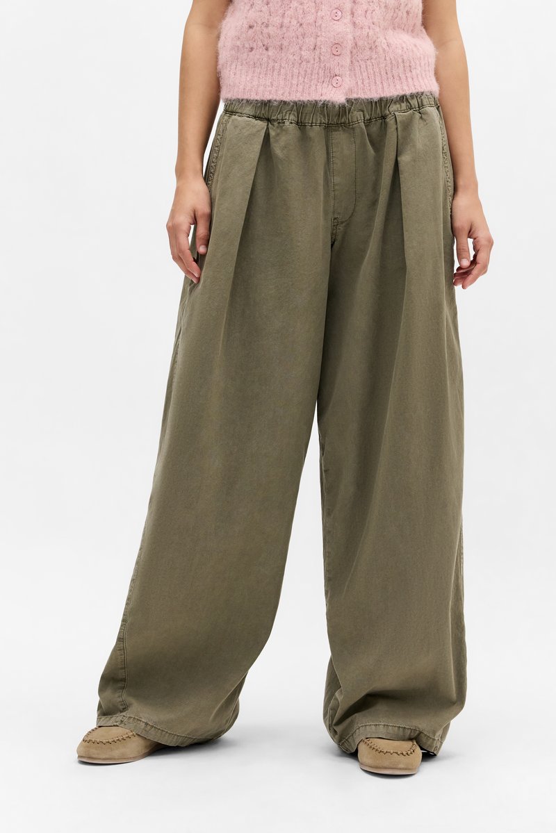 Pantalones de pierna ancha color caqui con un diseño plisado, cinturilla elástica y tela suave y texturizada; combinados con calzado estilo mocasín en color beige claro.