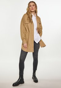 Oversize beżowa kurtka z dużymi kieszeniami noszona na białej koszuli, w połączeniu z ciemnymi skinny jeansami i czarnymi botkami za kostkę. Gładki materiał i swobodny styl.