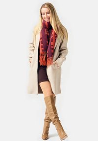 Long beige coat, maroon and orange patterned scarf with fringe, fitted brown dress, and knee-high smooth tan leather boots.