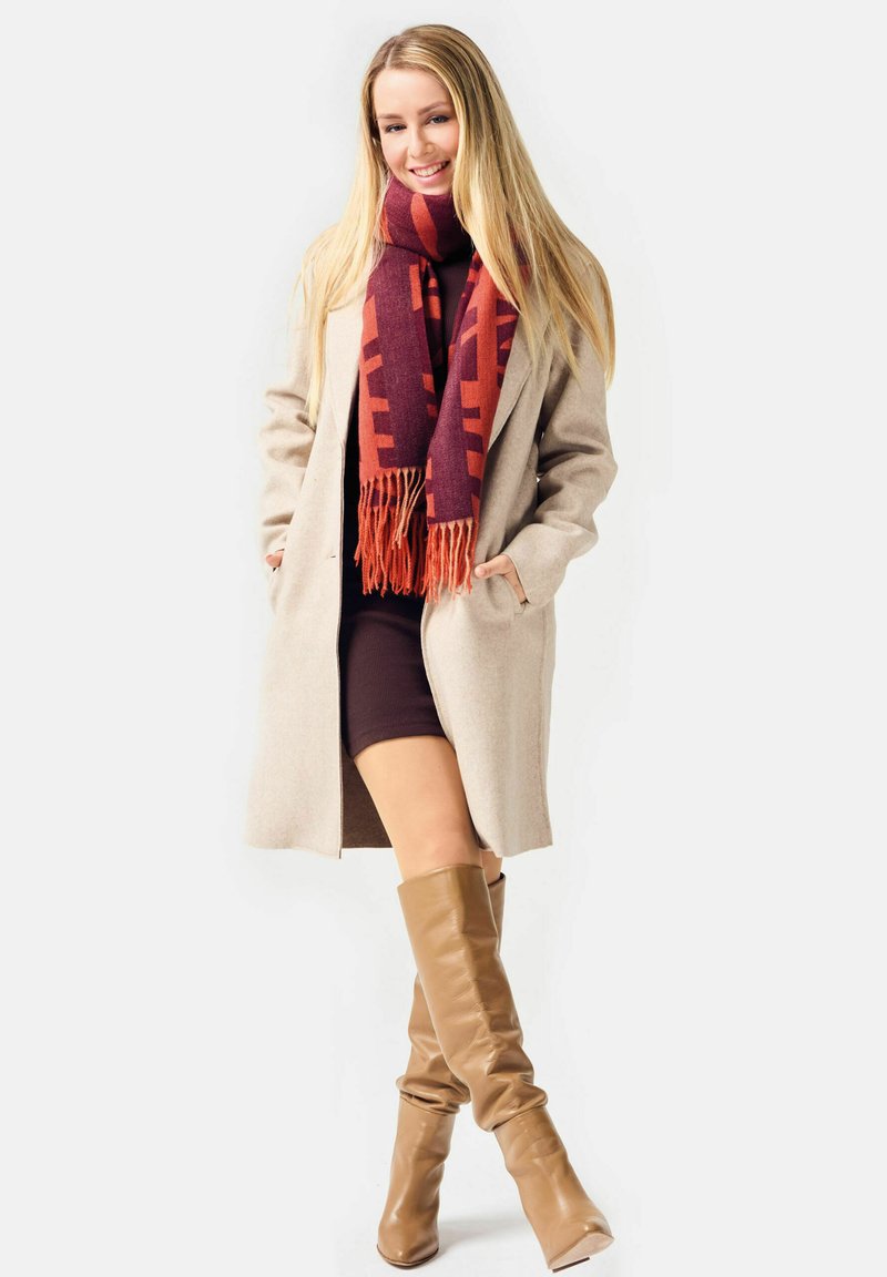 Long beige coat, maroon and orange patterned scarf with fringe, fitted brown dress, and knee-high smooth tan leather boots.