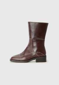 Bottine en cuir marron à bout carré et à talon bas, avec des coutures contrastées le long des seams. Fond clair.