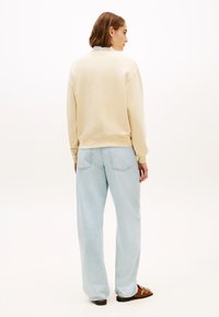 Ανοιχτό μπεζ φούτερ με ριμπ μ ανσέτες, συνδυασμένο με φαρδιά ανοιχτό μπλε τζιν. Το μοντέλο στέκεται με την πλάτη, φοράει καφέ σανδάλια.