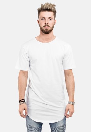 Fehér, rövid ujjú póló kerek nyakkivágással, egyenes vonalú fazonnal és hosszú aljjal, világoskék, testhez simuló farmernadrággal viselve.
