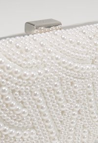 Vit pärlbehandlad kuvertväska med en strukturerad yta, som har små och stora runda pärlor samt ett metallspänne högst upp. Förstärkta kanter är synliga.