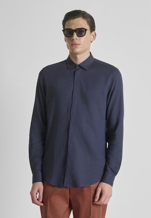 Man met zwarte zonnebril, donkerblauw overhemd met knopen en lange mouwen, en roestbruine broek tegen een effen grijze achtergrond.