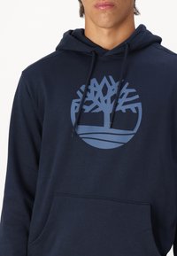 Navyblauwe hoodie in een zachte stof, met een groot lichtblauw boompje-logo en een centrale zak. De koordjes zijn zwart met metalen uiteinden.