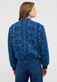 Kobieta zwrócona w prawo, ubrana w niebieską wzorzystą kurtkę pikowaną i ciemnoniebieskie dżinsy, na jednolitym tle.