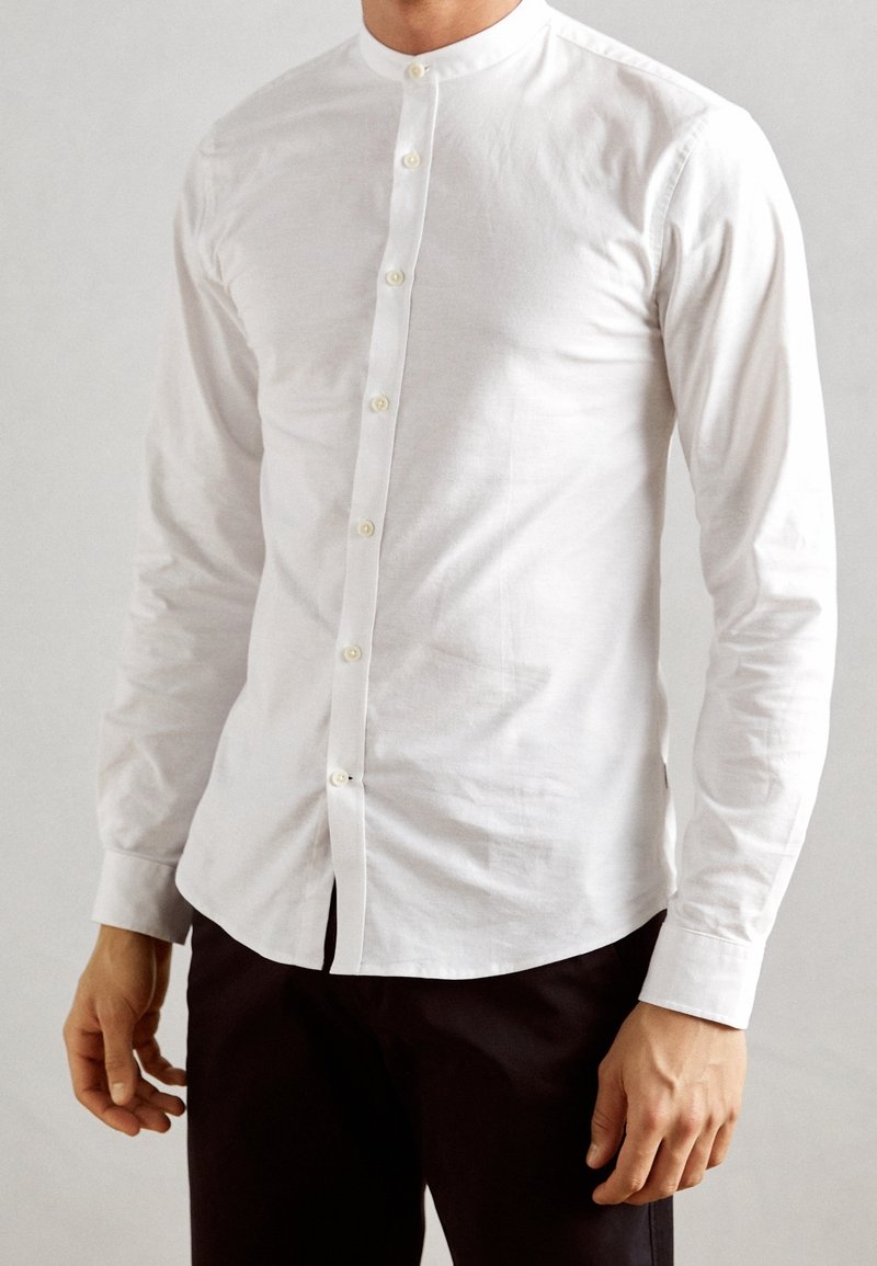 Homme portant une chemise blanche cintrée à manches longues avec un col mandarin et un pantalon noir, debout devant un fond uni.