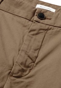 Pantaloni di tessuto marrone con chiusura a bottone, cuciture visibili e una texture leggermente stropicciata. L'etichetta riporta "hessnatur."