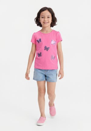 Ung jente går, iført en rosa kortermet skjorte med paljett sommerfugler, lyse denimshorts og rosa slip-on sko.