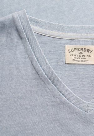 Lyseblå V-hals t-shirt med synlig syet halsudskæring og en beige mærkat med teksten "Superdry Co. Craft & Detail Trade Mark British Design."