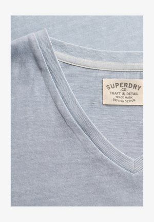 Jasnoniebieska koszulka z dekoltem w serek, z widocznym przeszytym dekoltem i beżową metką z napisem „Superdry Co. Craft & Detail Trade Mark British Design.”