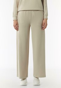 Beżowe szerokie spodnie dresowe z miękkiego materiału, z elastycznym paskiem w talii i luźnym krojem, w połączeniu z białymi butami sportowymi.