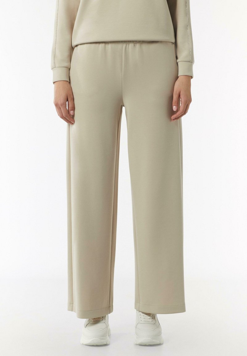 Beżowe szerokie spodnie dresowe z miękkiego materiału, z elastycznym paskiem w talii i luźnym krojem, w połączeniu z białymi butami sportowymi.