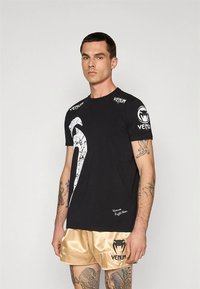 Melna īsroku T-krekls ar lielu baltu plaisājošu logotipu, Venum zīmolu uz pleciem un šauru piegriezumu.