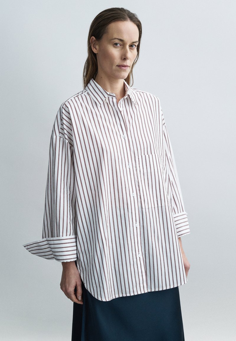 Pruhovaná košeľa v bielej a bordovej farbe, vyrobená z ľahkej látky. Má golierový výstrih, zapínanie na gombíky a nadmerné rukávy.