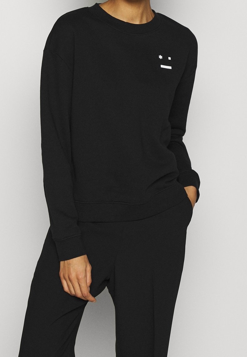 Personne portant un sweat-shirt noir à manches longues et un pantalon noir assorti, avec un petit motif blanc près de l'épaule gauche.