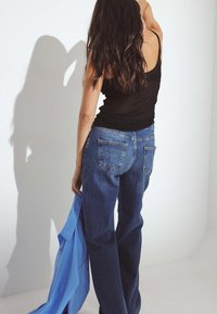 Tamsus tank topas, mėlynos džinsinės plačios kelnės su laisvu modeliu ir šviesiai mėlynas marškinys, laikomas vienoje rankoje, prieš paprastą šviesią fono.