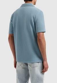 Korte mouwen polo-shirt in lichtblauw, gemaakt van textuurstof, met een klassieke kraag en een losse pasvorm. Er worden denim jeans gedragen.