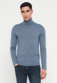 Mann trägt einen hellblauen Rollkragenpullover und dunkle Jeans, steht mit einer Hand in der Tasche vor einem schlichten weißen Hintergrund.