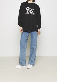 Sudadera negra con texto gráfico en blanco que dice "THE MAP ERA 67/21 TOUR", combinada con jeans de pierna ancha de color azul claro y zapatillas blancas.
