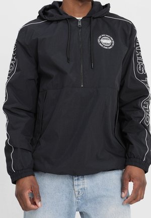 Felpa nera con cappuccio e cerniera frontale, logo circolare sul petto, testo bianco sulle maniche, indossata con jeans azzurri chiari.