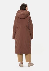 Manteau long marron avec capuche, fabriqué à partir d'un matériau lisse et imperméable. Possède des poches latérales et une coupe décontractée. Porté par-dessus un pantalon beige.