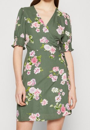 Kvinde iført en grøn slå-om-kjole med rosa og hvide blomsterprint, puffede ærmer med smockede manchetter og en V-hals.