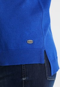 Jasnoniebieski sweter z dzianiny z małą owalną metalową metką nad bocznym rozcięciem, połączony z ciemnoniebieskimi dżinsami.