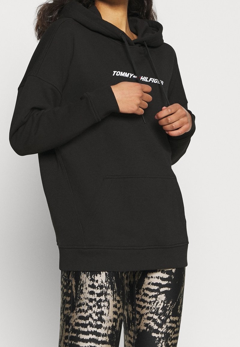 Felpa nera con cappuccio e tasca frontale, con il testo bianco "TOMMY HILFIGER". Abbinate a leggings lucidi con motivo a serpente.