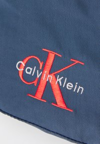 Tengerkék szövet finom textúrával, amelyen hímzett "Calvin Klein" logó látható; a "CK" élénk piros színű, míg az "alvin Klein" fehérben.