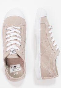 Sneakers in tela beige con punte e suole in gomma bianca, dotate di lacci e cuciture bianche. Include un'etichetta con il logo all'interno.