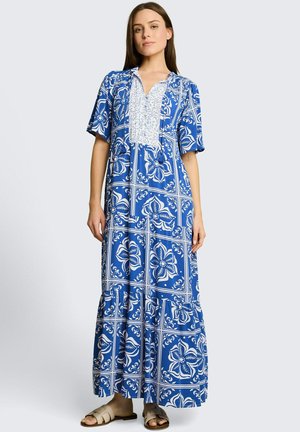 Femme portant une robe longue à motif floral bleu et blanc, manches courtes et sandales plates beiges, debout devant un fond uni.