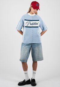Personne portant une casquette rouge, un haut zippé bleu clair à motifs avec le texte « Interdit », un short en jean, des chaussettes blanches et des mocassins noirs, debout les mains dans le dos.