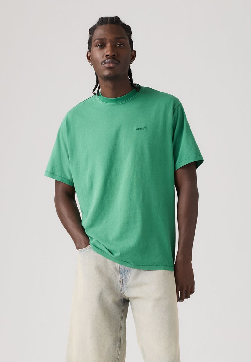 Jeune homme aux cheveux tressés portant un t-shirt vert Levi's et un jean clair, debout avec une main dans la poche devant un fond uni.