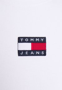 Tejido de algodón blanco con un parche bordado rectangular. El parche tiene "TOMMY" en la parte superior, "JEANS" en la parte inferior, con colores rojo, blanco y marino.