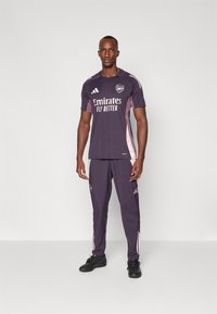 Ensemble de sport violet avec un t-shirt à manches courtes et un pantalon long. Présente des accents blancs, un logo et un design à col rond. Porté avec des chaussures noires.