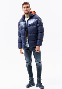 Marine pufferjack met een glanzende afwerking, oranje voering van de capuchon, ritssluiting, gecombineerd met versleten denim en donkerblauwe hoge sneakers.