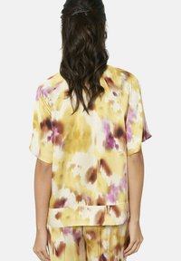 Bluzka z krótkim rękawem z jedwabistego materiału, w żółtej bazie z abstrakcyjnymi fioletowymi i brązowymi wzorami, luźny krój i prosty dół.