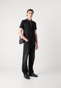 Svart pikétröja med grå krage, i kombination med mörk denimjeans. Modellen håller en liten svart axelväska med en blå logotyp.